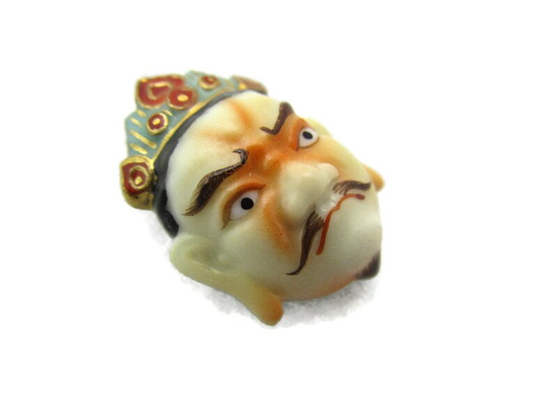 May include: A porcelain figurine of a man's face with a crown. The face is painted with details, including a mustache, eyebrows, and eyes. The crown is painted in red, blue, and gold.