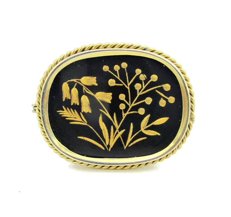 Puede incluir: Un broche dorado con fondo de esmalte negro que presenta un dise&ntilde;o floral detallado en oro. Las flores son estilizadas y tienen un aspecto delicado e intrincado.