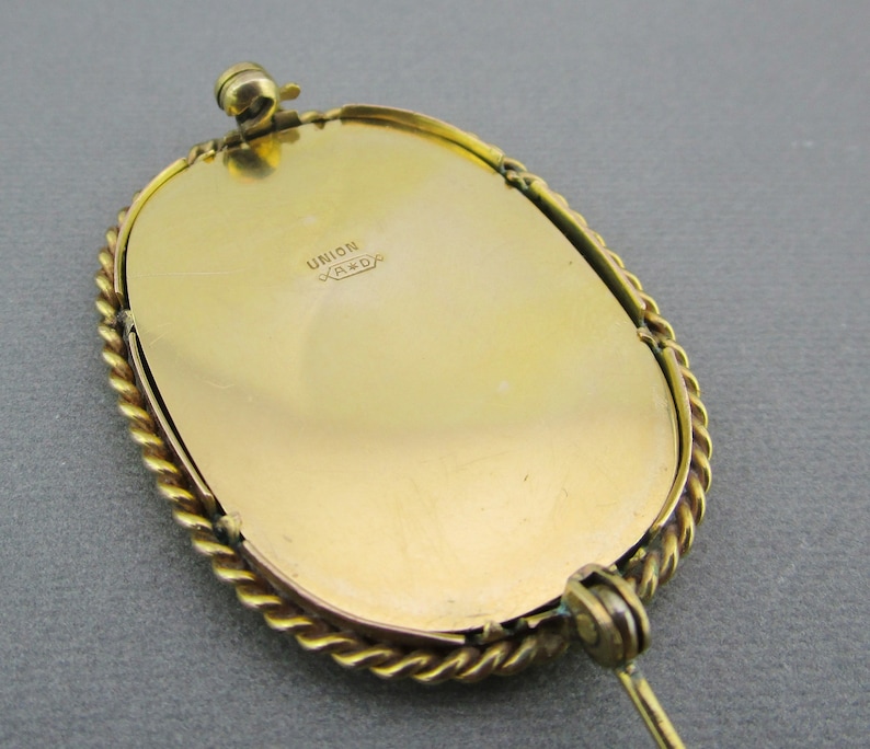 Puede incluir: Broche ovalada dorada con un borde de cuerda y un pasador en la parte posterior. La parte posterior de la broche est&aacute; grabada con el texto "UNION X*A*D*"