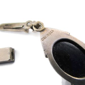 Puede incluir: Primer plano de un colgante de plata con una piedra ovalada negra. El colgante tiene un cierre y est&aacute; marcado con "STERLING" en la parte posterior.