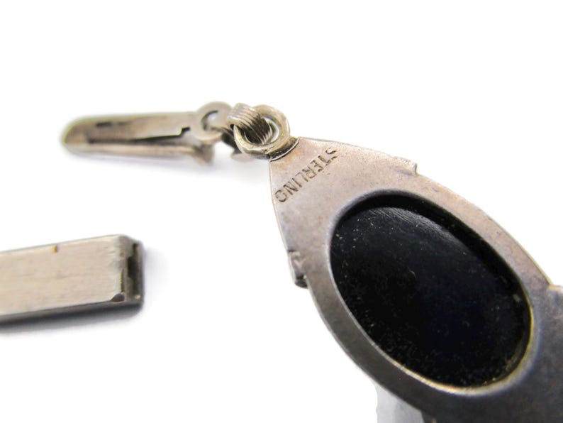 Puede incluir: Primer plano de un colgante de plata con una piedra ovalada negra. El colgante tiene un cierre y est&aacute; marcado con "STERLING" en la parte posterior.