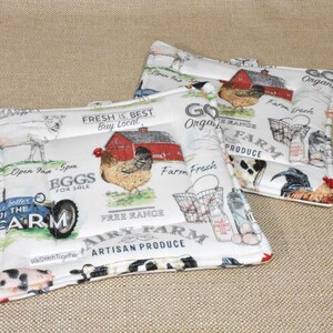 May include: Two white cotton pot holders with a farm theme. The pot holders feature a red barn, chickens, cows, a tractor, and the text "Fresh is Best Buy Local", "Free Range", "Dairy Farm Artisan Produce", "Go Organic", "Farm Fresh", and "Farm Produce".