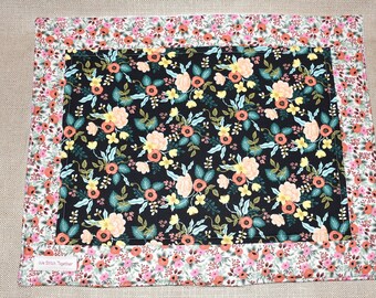 May include: A black floral fabric placemat with a pink and orange floral border. The placemat has the text "We Stitch Together" printed on the bottom edge.