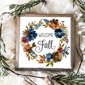 May include: Square wooden sign with a watercolor floral wreath in shades of blue, orange, and red, with the words "WELCOME Fall" in black script. The sign is surrounded by greenery, pinecones, and a white fluffy background.