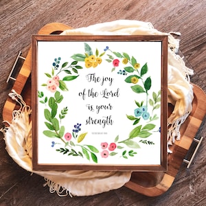 May include: Square wooden framed art print featuring a watercolor floral wreath in shades of pink, yellow, and blue. The text reads "The joy of the Lord is your strength" from Nehemiah 8:10. The print is displayed on a wooden tray.