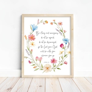 May include: A framed print with a light wood frame. The print features a floral wreath in watercolor with text that reads "Be strong and courageous, do not be afraid, do not be discouraged..."