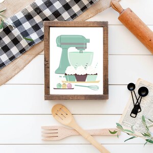 May include: A framed illustration of a green stand mixer with a bowl and cupcakes. The cupcakes are decorated with sprinkles and frosting. There are also some colorful sprinkles on the table.