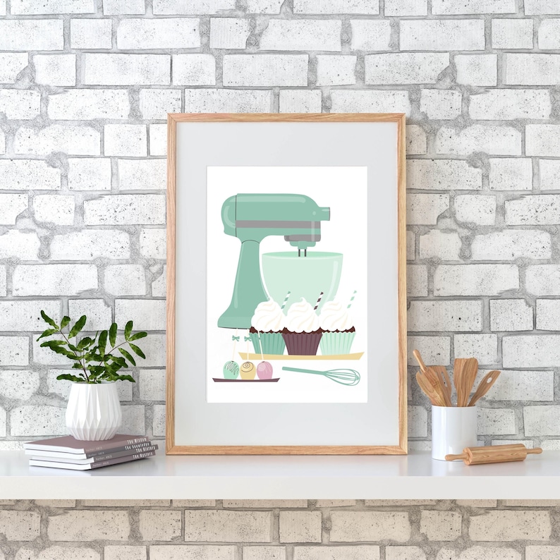 May include: A framed print of a green stand mixer with a bowl, three cupcakes with frosting and sprinkles, and a tray of chocolate and pink cake pops. The print is on a white background and has a wooden frame.