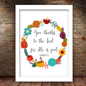 May include: A white framed print with a floral wreath design. The wreath is made up of colorful flowers in shades of red, orange, yellow, blue, and green. The text inside the wreath reads "Give thanks to the Lord for He is good. 1 Chronicles 16:34".