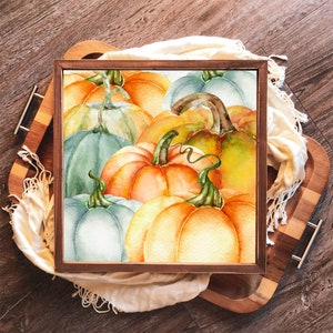 May include: A square wooden framed artwork featuring a watercolor painting of pumpkins in shades of orange, green, and blue. The pumpkins are clustered together, with visible stems and vines. The artwork is displayed on a wooden tray with a white scarf.