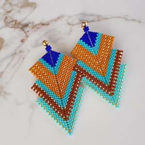 Könnte beinhalten: Ein Paar geometrische Ohrringe mit einem mehrschichtigen Design. Die Ohrringe sind aus blauen, türkisfarbenen, orangefarbenen und braunen Perlen gefertigt. Die Ohrringe sind an goldfarbenen Haken befestigt.