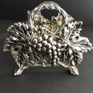 May include: A silver-toned metal napkin holder featuring a detailed relief design of grape clusters and vine leaves. The structure includes a central arched handle and four small feet, with an openwork pattern throughout the metallic surface.