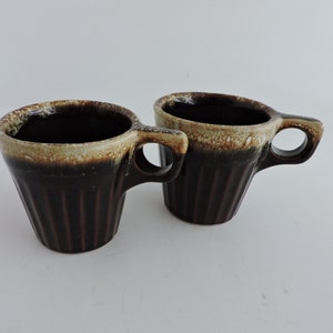May include: Two brown ceramic mugs with a ribbed design. The rims are a light tan color, and the handles have a circular opening. The interior of the mugs is dark brown. The mugs are set against a white background.