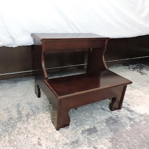 Puede incluir: Un taburete de madera de dos escalones con acabado en cerezo oscuro. El taburete tiene un escalón superior e inferior rectangular con soportes laterales curvos. El taburete está colocado sobre una alfombra estampada cerca de una cama.