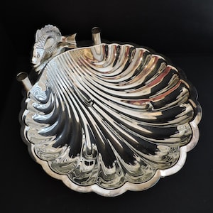May include: A polished silver-toned metal dish shaped like a large scallop shell. The dish features a decorative fish figure attached to one side and is supported by three small cylindrical feet, creating a reflective and textured surface.