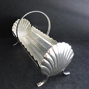 May include: A silver-colored, shell-shaped basket with a handle. The basket has a rectangular, perforated body and is supported by three curved feet. The ends of the basket are shaped like scallop shells.