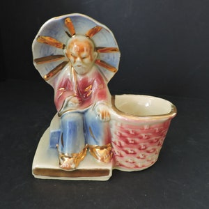 May include: A vintage ceramic figurine of an Asian man seated with a basket and a sun-like halo. The figurine features a color palette of red, blue, gold, and cream. The basket has a woven texture, and the man is wearing a red robe and blue pants.