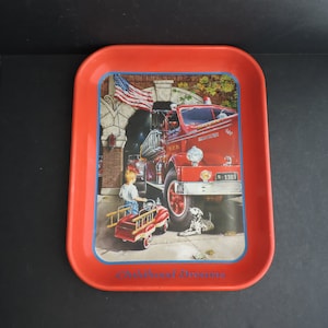 Puede incluir: Bandeja rectangular roja de metal con borde azul. La bandeja presenta una colorida ilustración de un camión de bomberos rojo vintage, un niño, un perro dálmata y una bandera estadounidense. Las palabras "Childhood Dreams" están impresas.