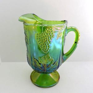 May include: Green iridescent glass pitcher with a handle and spout. The pitcher features embossed grape and leaf designs. The base is round and the pitcher has a curved handle. The glass has a shiny, reflective surface.
