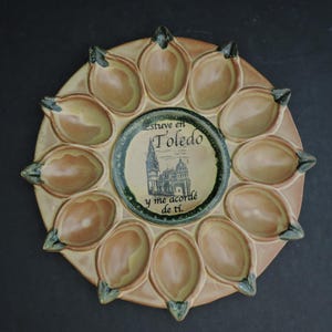 Könnte beinhalten: Keramik-Servierplatte mit floralem Design. Die Platte hat einen zentralen kreisförmigen Bereich mit Text und einer Gebäudeillustration, umgeben von zwölf blütenblattförmigen Fächern. Die Farben sind Beige, Grün und Braun.