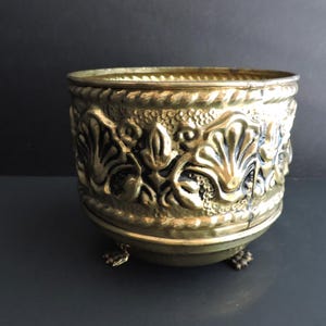May include: A round brass planter featuring an embossed shell and leaf pattern around the exterior. The vessel has a rope-style border at the top rim and rests on three small, detailed claw-shaped feet. The metal surface shows a polished finish.