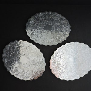 May include: Three circular silver-toned metal coasters featuring a scalloped edge and an intricate embossed scrollwork pattern across the surface. Each coaster has a smooth, reflective finish with a plain center surrounded by detailed floral motifs.