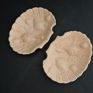 Pode incluir: Duas travessas em forma de concha na cor pêssego claro. As travessas têm um design em relevo detalhado de conchas e algas marinhas. As bordas das travessas são onduladas. Em um fundo escuro.