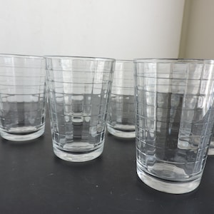 May include: A set of five clear glass tumblers, each featuring a distinct grid pattern of thin horizontal and vertical lines etched into the exterior surface. The drinking glasses have a cylindrical shape with a flat base and a smooth, transparent finish.