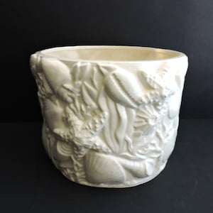 May include: A cylindrical off-white ceramic planter featuring a raised, textured relief pattern of various seashells, starfish, and seaweed wrapping around the entire exterior surface of the vessel with a smooth, glossy finish.