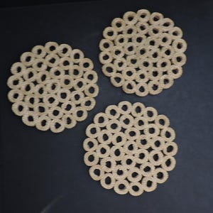 Puede incluir: Tres posavasos circulares construidos con cuerda de fibra natural de color tostado claro enrollada. Cada posavasos presenta un patrón calado repetitivo de bucles interconectados, creando una superficie texturizada con un diseño uniforme.