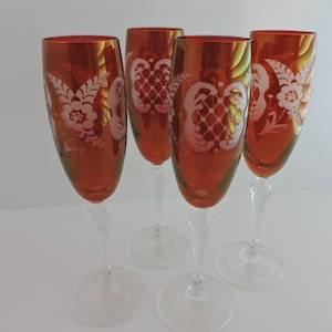May include: A set of four tall champagne flutes featuring deep amber-colored glass bowls with etched white floral and lattice patterns. Each glass has a clear, slender stem and a circular base, standing upright in a group of four distinct pieces.