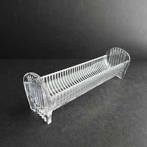 May include: A clear, rectangular glass dish with a ribbed design. The dish has curved ends and sits on small feet. The interior features vertical ridges, creating a decorative and functional piece for serving or display.