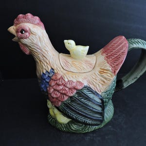 Könnte beinhalten: Eine Keramik-Teekanne in Form eines Hahns, mit einem kleinen Küken auf dem Deckel. Der Hahn ist in Beige-, Rot-, Blau- und Grüntönen bemalt. Der Griff und der Sockel sind grün. Ein dekorativer Küchenartikel.