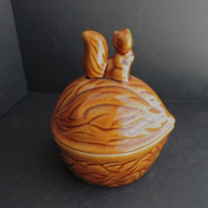 May include: A ceramic nut-shaped container with a squirrel figurine on top. The container is a warm brown color with detailed texture, and the squirrel is a lighter shade of brown. The lid fits snugly, suggesting it's a storage item.