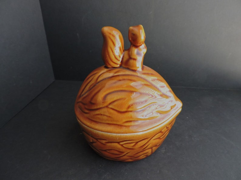 May include: A ceramic nut-shaped container with a squirrel figurine on top. The container is a warm brown color with detailed texture, and the squirrel is a lighter shade of brown. The lid fits snugly, suggesting it's a storage item.