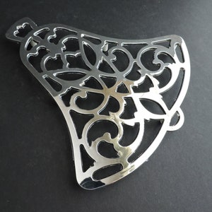 May include: A silver-colored, decorative metal trivet with an intricate, openwork design. The trivet is shaped like a bell and features swirling, floral-inspired patterns. It is designed to protect surfaces from hot cookware.