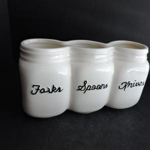 May include: A white ceramic utensil holder with three connected compartments. Each section is shaped like a mason jar and labeled with black cursive text: "Forks", "Spoons", and "Knives". The holder is designed to organize kitchen utensils.