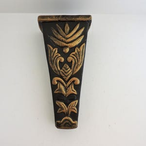 May include: A black and gold decorative wall bracket with ornate floral and leaf designs. The bracket has a triangular shape and is mounted on a white wall. The gold accents highlight the intricate details of the carved patterns.
