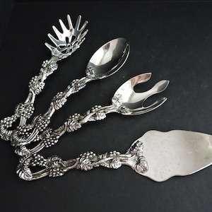 Könnte beinhalten: Silbernes Servierbesteck-Set auf schwarzem Untergrund. Das Set umfasst eine Salatgabel, einen Löffel, eine Serviergabel und einen Tortenheber. Die Griffe sind mit einem Weinreben-Design verziert.