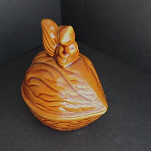 May include: A ceramic container shaped like a walnut, with a lid featuring a stylized nut design. The container is a warm brown color with glossy finish. The lid has a detailed, textured surface.