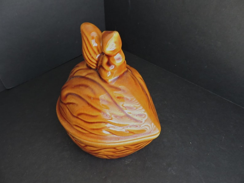 May include: A ceramic container shaped like a walnut, with a lid featuring a stylized nut design. The container is a warm brown color with glossy finish. The lid has a detailed, textured surface.