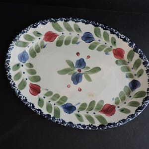 Puede incluir: Plato ovalado de cerámica con un diseño floral pintado a mano. El plato blanco presenta un borde de hojas azules y verdes, flores rojas y azules y un borde festoneado azul oscuro. También hay un motivo floral central.