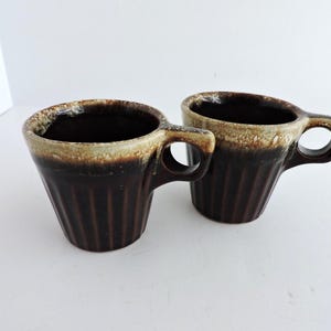 Pode incluir: Duas canecas a condizer com um corpo castanho escuro canelado e um esmalte de gotejamento em tom creme que contrasta na borda superior. Cada caneca tem uma pega circular e uma abertura larga, exibindo um acabamento brilhante de dois tons.