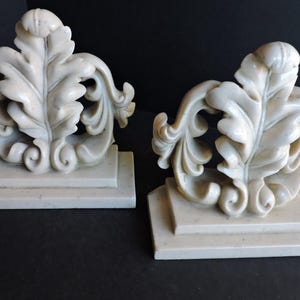 Puede incluir: Par de sujetalibros en forma de hoja, de color blanquecino. Cada sujetalibros presenta un diseño de hoja tallada con detalles y remolinos, montado sobre una base rectangular. Fabricados en un material sólido, posiblemente similar a la piedra.