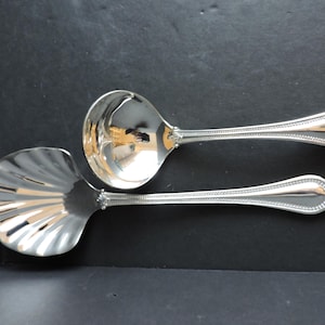 May include: Two silver serving utensils on a black surface. One is a ladle with a round bowl and a long handle. The other is a slotted serving spoon with a shell-shaped bowl and a long handle. Both have decorative detailing.