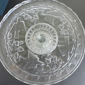 Puede incluir: Un soporte para pasteles de vidrio transparente con un patrón en relieve de pájaros y racimos de fresas alrededor del plato circular. El centro del soporte tiene un diseño de celosía texturizado y el borde exterior tiene un acabado festoneado.
