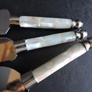 May include: Close-up of three butter knives with mother-of-pearl handles and silver-colored metal blades and accents. The handles are made of rectangular pieces of mother-of-pearl. The blades are flat and rectangular.