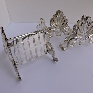 May include: Silver-colored metal serving dish with a decorative, ornate design. The dish has a rectangular basket shape with vertical slots and is supported by decorative feet. Two additional decorative pieces are also visible.