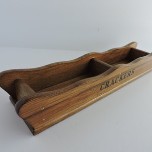 May include: A long, rectangular wooden serving tray with a dark brown finish and wavy side edges. The tray is divided into two sections by a central wooden partition and features the word CRACKERS printed in black lettering on the front side.