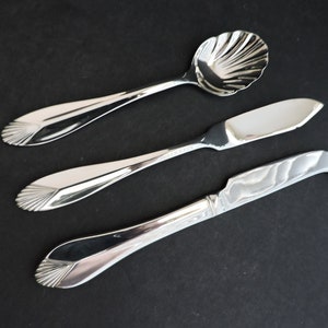 May include: Three silver serving utensils on a black surface. The set includes a spoon with a shell-shaped bowl, a butter knife, and a cheese knife. Each piece has a decorative fan-shaped design at the handle's end.